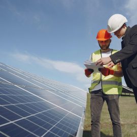 Low angle of young inspector and foreman in hardhats checking documentation against modern solar panels in field